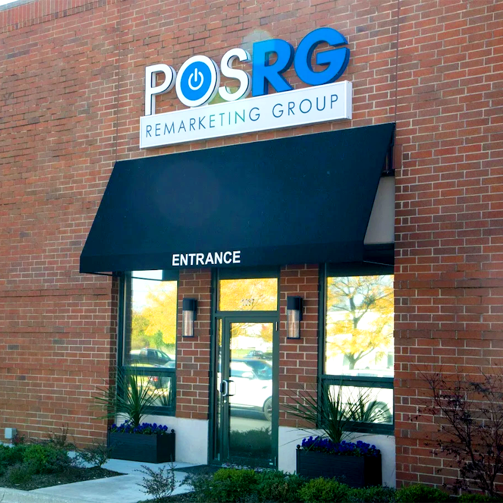 POSRG Remarketing Group storefront featuring a red brick exterior, a large logo sign with a blue power button icon above the black awning labeled “ENTRANCE,” and landscaped planters with greenery and flowers.
