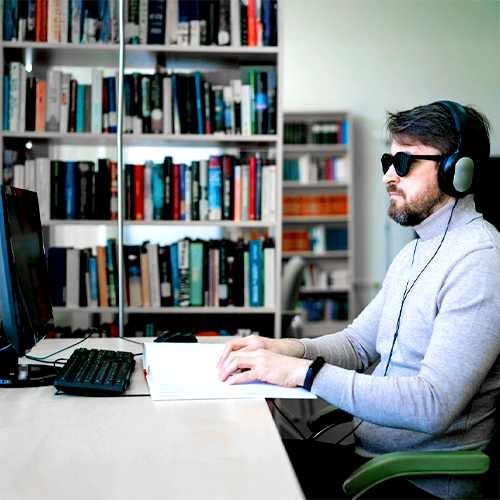 A visually impaired man wearing headphones and sunglasses reads braille at a desk with a computer monitor, in front of a wall of bookshelves in a well-lit library or office setting.