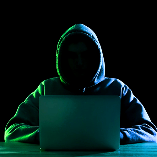 Silhouetted figure in a hoodie sitting in front of a laptop in a dark room, symbolizing cybersecurity threats or hacker activity, with green lighting accentuating the shadows.