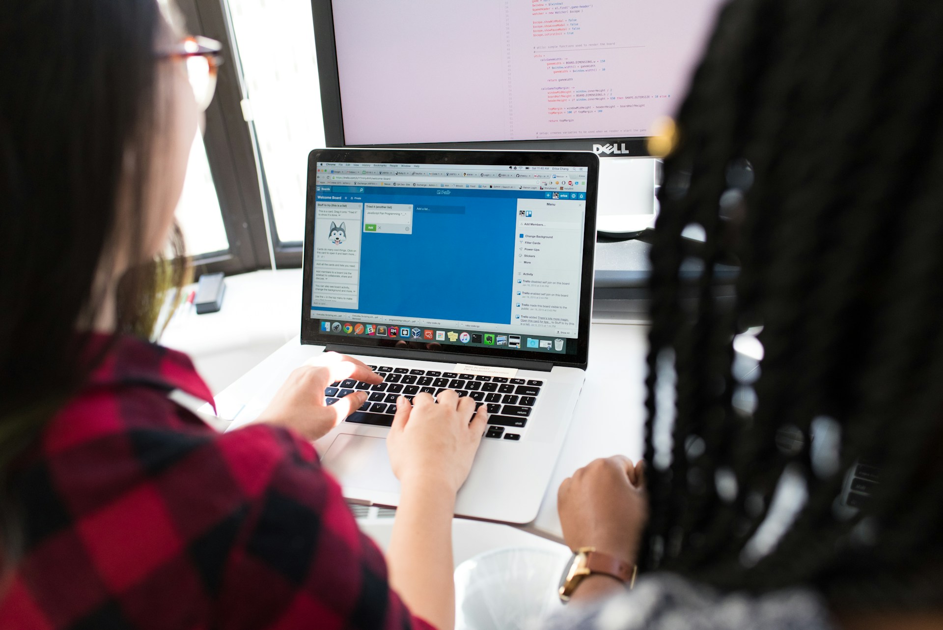 Two people work together at a desk, one typing on a laptop displaying a project management tool, with another computer monitor visible in the background.